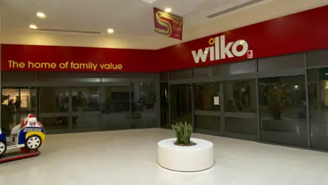 The shop frontage of the old Wilko store in Weston Favel - a black glass front with a red sign and a child's police ride in front and a small decorative plant at the entrance.