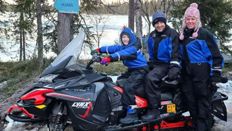 Wayne O'Neil and his son Finn sitting on a snowmobile with Lisa O'Neil standing next to them. They are all wearing snow suits and woolly hats, but there is no snow on the ground around them. There is a lake in the background.