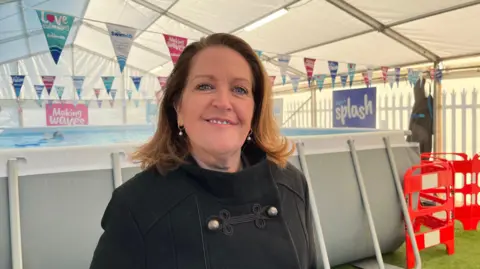 Tom Jackson/BBC A woman wearing a black coat with gold buttons, standing in front of the pop-up pool, smiling for the camera.