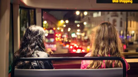 Getty Images the back of two young women's head. they are sitting on a bus at night looking onto traffic