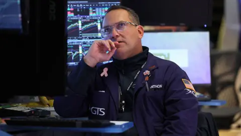 Getty Images A trader in a dark blue coat with an American flag on his sleeve sits near computers at the New York Stock Exchange in October 2025, his eyes lifted to the screens above him.