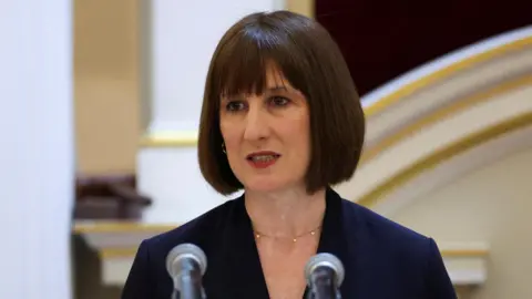 Rachel Reeves, a woman with a dark bob hair style, and a blue suit, stands in front of two microphones