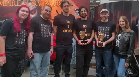 Six people from the team behind the game Vampire Survivors, standing up in front of a banner for the game Vampire Survivors at the video game conference WASD, two of them are holding BAFTA awards.