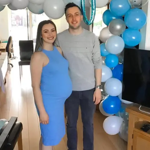 Kianty and Conor Simpson Kianty in a blue dress with a visible baby bump is smiling next to Conor. There's a table of buffet food in the background and a balloon arch with white and pale and dark blue balloons. A metallic blue balloon at the top of the arch reads ''Baby"