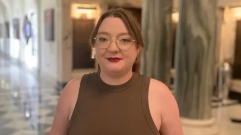 A woman in a brown vest is looking at the camera. She wears glasses and red lipstick. Behind her are marble pillars and an arched hallway. 