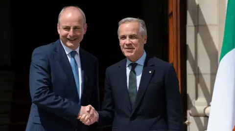Martin and Carnety shake hands and a tri-colour hands to the right hand side of the pair. Martin a bit taller and bald on top. Carney has short grey hair. 