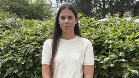 Ella has long dark hair and wears a cream coloured top and stands in front of a hedge.