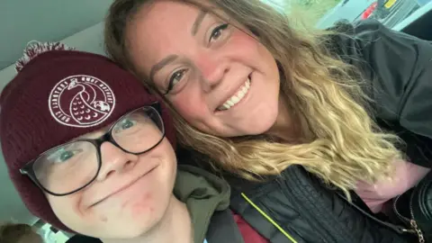 Harry Basham and his mum Leanne Golding are smiling at the camera. They are sitting inside a car. Harry is wearing a burgundy hat with Taunton Town Football Club written on it. Leanne is wearing a jacket and a pink top. She has long wavy hair.