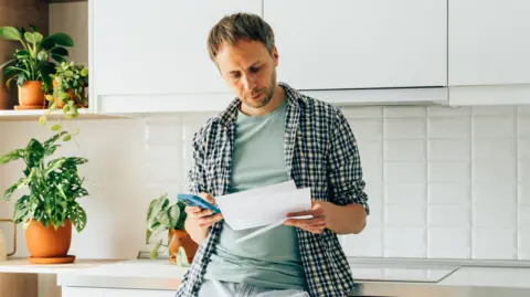 Man leans against a work surface in a kitchen holding paperwork and a phone.