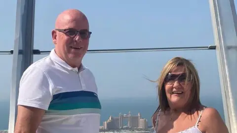 Steven Stewart Steven, a tall bald man wearing sunglasses standing next to his wife Marie who has blonde hair and is also wearing sunglasses. They are standing a height with a grand building visible in the background.