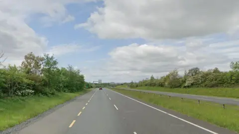 An empty road with grass and trees on either side. There is a blue sky above with some clouds. 