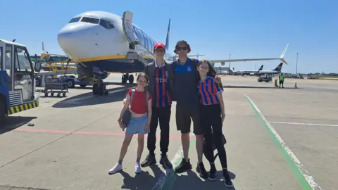 Jimmy Joyce Four people stand together on an airport runway in front of a Ryanair plane with its door open and steps in place, with ground vehicles and other aircraft visible in the background under a clear blue sky.