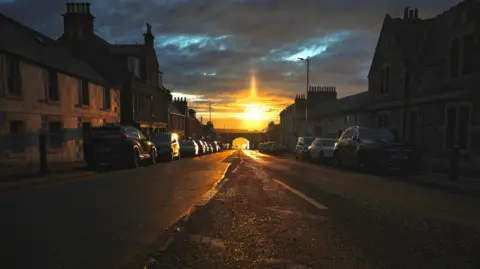 Pamela McKay A dark village street with cars parked along the pavements. There is bridge with an archway at the end of the road. The sunset reflects orange along the street and the sky is dark.