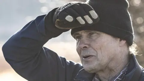 Countryside Regeneration Trust Close up of Dr Vince Lea wearing a black beanie hat, black gloves and a jacket. He is outside and has one hand against his forehead shielding his eyes from the sun as he looks to away from the camera.
