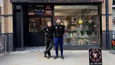 Image of a man with his arm around the shoulders of another man in front of their shop. There are collectable toys in the shop window.
