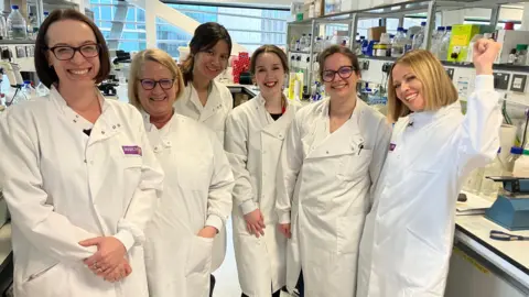 Kimberley Walsh is standing next to five scientists, who are all wearing white lab coats, in a laboratory. 