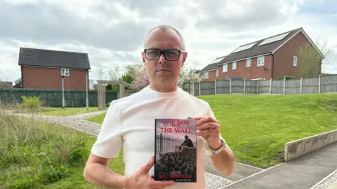 GRAEME BAMFIELD Graeme, a man in his 50s, he wears glasses and looks stoically at the camera with his book called the boy over the wall, which has an image of a boy being pulled over a wall.