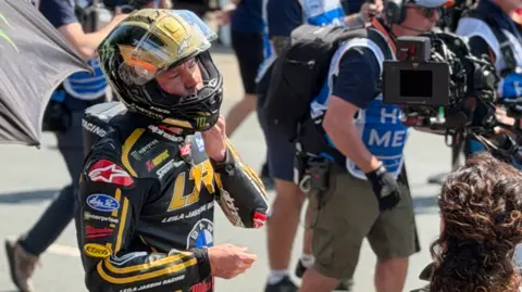 Frank Schuengel Channing Tatum is wearing black and gold leathers and a matching helmet. There are people walking behind him and a film camera facing him to the right.