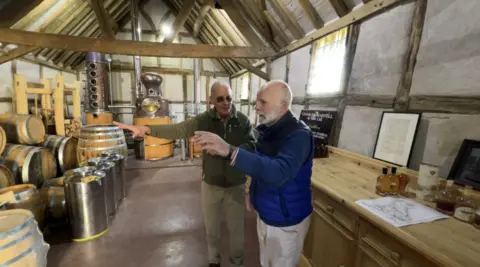 Charles Martell has grey hair and a beard and is wearing a blue jacket and Andrew Snell is wearing a khaki jumper. They are both wearing light trousers and are in a distillery room style room with a wooden desk in it.