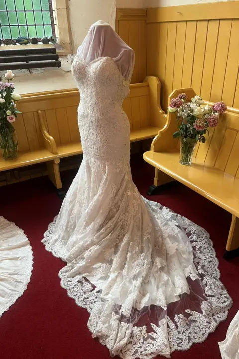 A wedding dress on a mannequin with church wooden seats behind it.