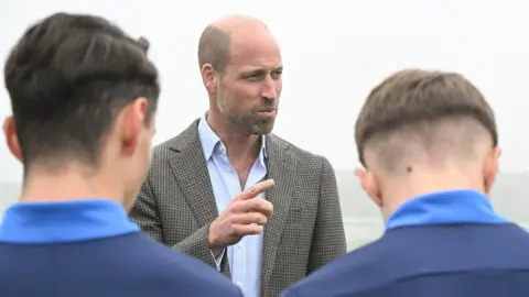 Price William is pointing a finger and two young men are looking at him with their backs to the camera. Prince William is wearing a pale blue shirt and a checked jacket. He has pale grey hair and a beard. It is a foggy day.