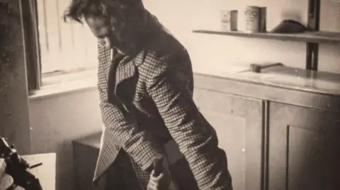 Jeff Towns Dylans Bookstore Dylan Thomas in a black and white photo standing beside the same window, except he has the legs of a chair in his hands and is twisting them with this arms.