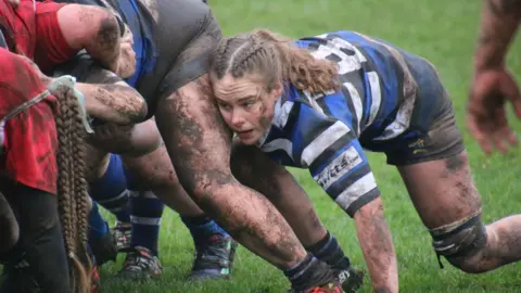 Dereck Thomson A female rugby player at the back of a scrum. She is knelt close to the floor. She is wearing a blue and white rugby kit and is covered in mud.