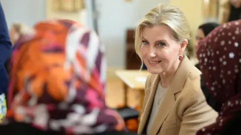 Kings Arms Project The Duchess of Edingburgh visiting the English Language club in April. Her blonde hair is pulled pack with a tendril at the front of her face, She is wearing a light coloured knit with a camel coat and dainty gold pendant