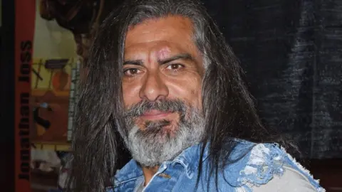 Jonathan Joss smiling for the camera with long black hair and a frayed denim shirt, pictured in 2021 