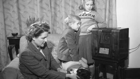 A black and white image of a woman and two children listening to a large wooden radio.