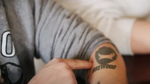 A man points to a Brewdog logo tattoo on his left arm