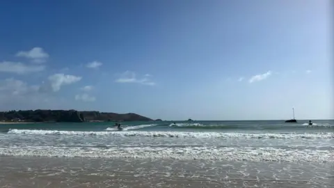 People in the water at St Brelade Bay. It is a sandy beach and there's a view of waves crashing on the sand. There's land in the distance on the left. The sky is blue with a few white clouds.