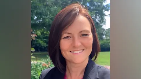 A woman with shoulder length smiles at the camera. She is wearing a black jacket over a pink top. She is standing in front of a tree on a sunny day.