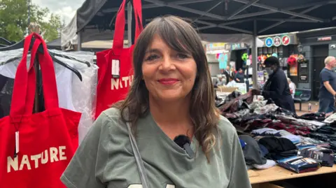 A woman smiles into the camera with a green top on and brown hair and red lipstick. Behind her, red fabric bags are hung up with the word "Nature" on them in white letters. On her other side, a table with folded clothes on can be seen.