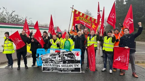 Eric Johnson/BBC A large group of protesters with red flags stand together outside the Vauxhall plant in Luton