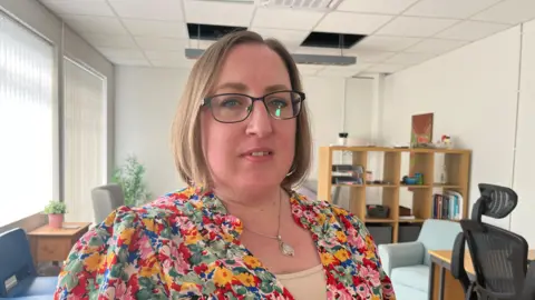 A woman with short brown hair and glasses wearing a floral colourful top and a silver pendant necklace