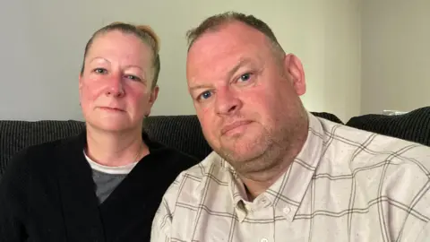 Lisa and Simon Adams sit on a sofa at home, looking upset. Mrs Adams is wearing a grey top and a black jumper and Mr Adams is wearing a cream shirt with a lined pattern.