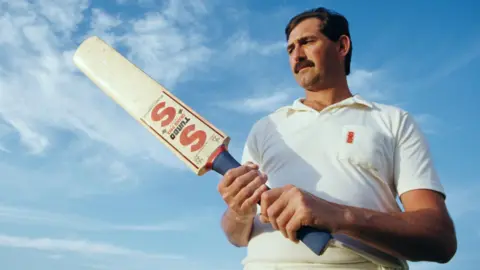 Getty Images Graham Gooch, wearing a white polo shirt, holds his bat up. He has brown hair and a moustache and it is a sunny day.
