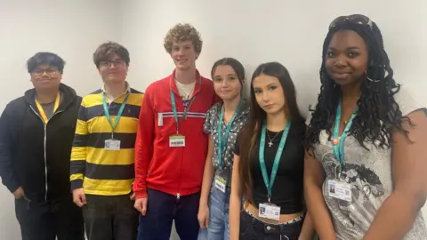 BBC/Grace Wood Three teenage boys and three teenage girls stand in a line against a white wall