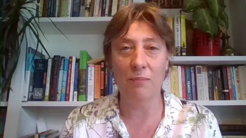 Head and shoulders image of Anni Byard who has short brown hair and is sitting in front of a bookcase