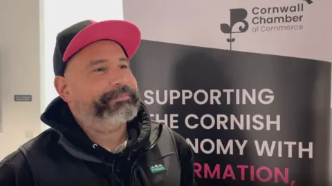 BBC A man wearing a hoodie and hat smiling, standing in front of a Cornwall Chamber of Commerce banner inside a white room.