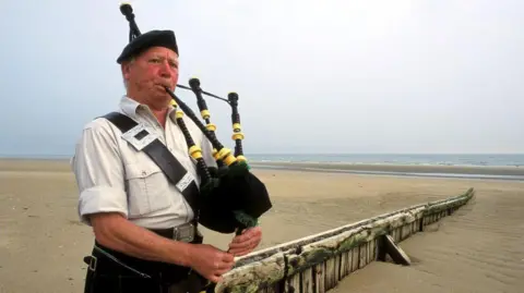 Getty Images Bill Millin berdiri di pantai. Dia bermain bagpipe. Bill mengenakan baret komando, rok dan kemeja berwarna terang dengan lengan bergulir.