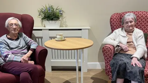 Two woman sit on large red-coloured chairs with a brown table between them, which has a cup of tea placed upon it, in a white room inside a care home