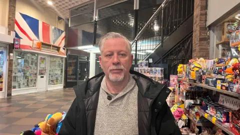 A man standing in a shopping arcade with toys for sale displayed behind him