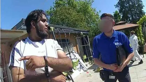 Essex Police A man in handcuffs and a white top. Next to him is a man from the RSPCA with his face blurred, wearing a blue top. They are standing outside. 