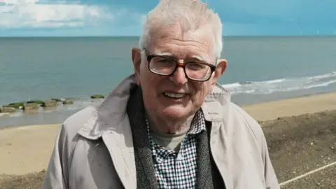 John Fairhall/BBC Andrew Fakes standing above a beach. He has white hair and is wearing glasses, a light coloured rain coat over a grey tweed jacket and checked shirt. Behind him is a sandy beach with seaweed-covered anti-tank blocks emerging from the bluey-green sea. Above it is blue sky.