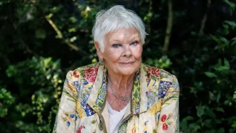 Dame Judi Dench in an florally embroidered jacket, sat in front of a background of green plants.