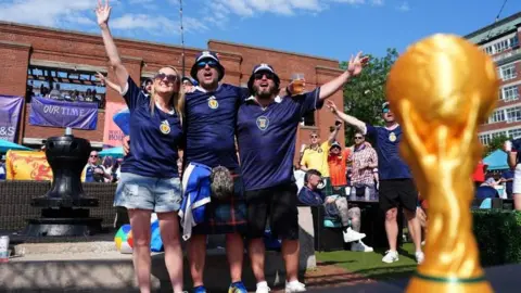 A group of Scotland fans cheering behind a gold World Cup trophy