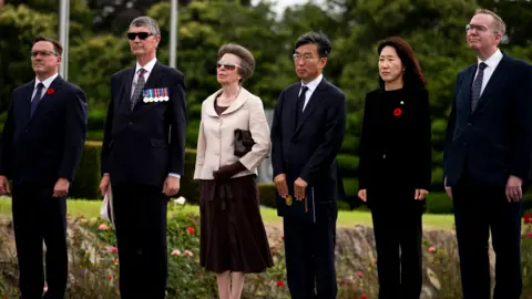 PA Media Princess Anne stood in a line with her husband, two other men and a woman at a memorial service. She has dark grey hair and is wearing a brown dress and cream jacket. Everyone is stood looking to the side to watch a flag raising ceremony.