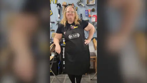 A woman with shoulder length strawberry blonde hair wears a black apron with the words 're-make Newport' on it stands with one hand on her hip infront of a blue wall full of tools.